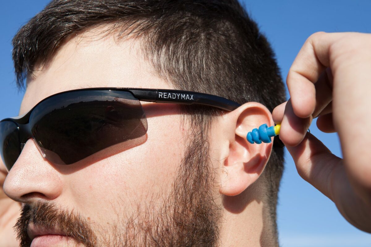 a man wearing soundshield safety glasses with retractable earplugs inserting the ear plugs into his ears.
